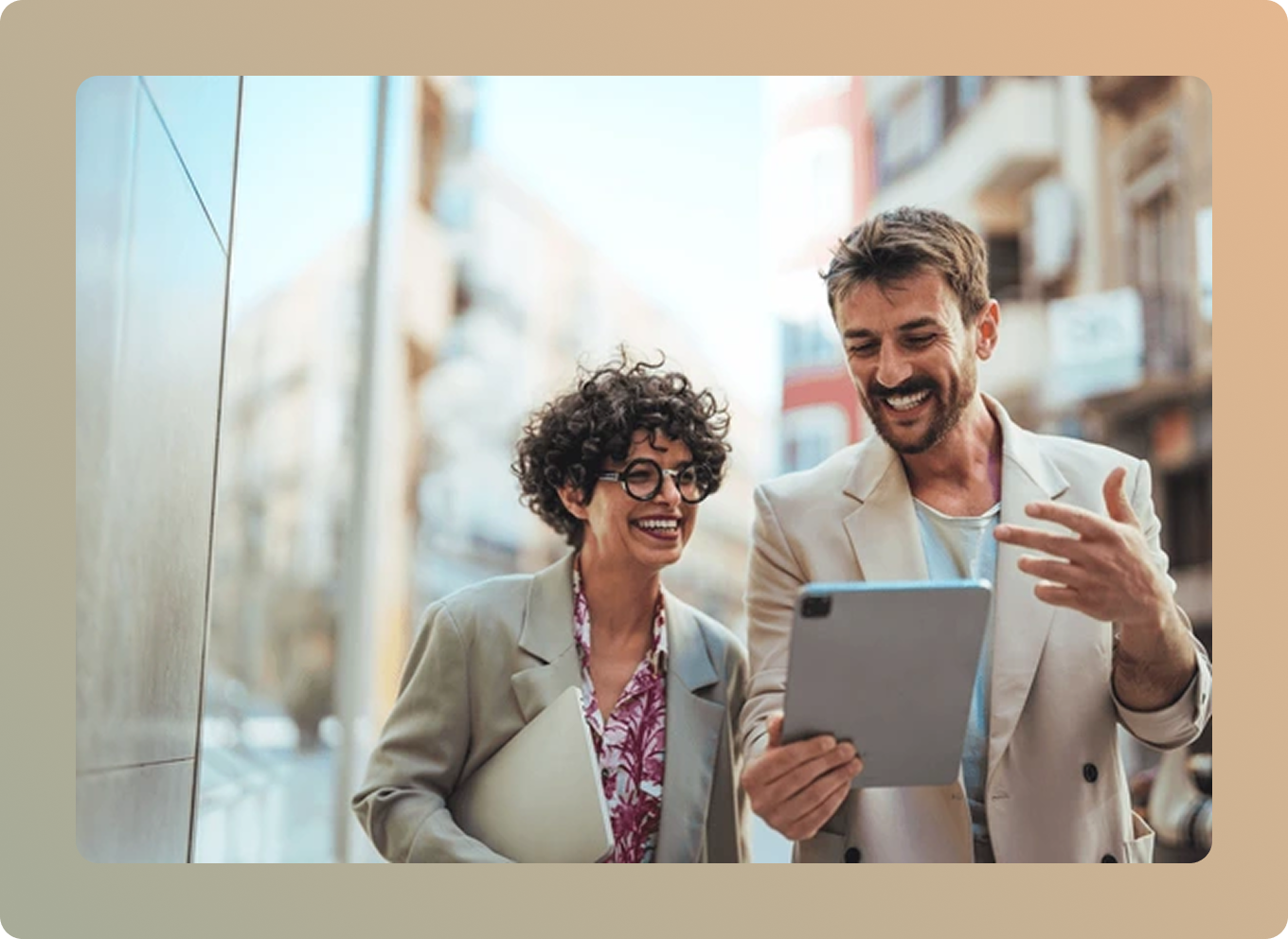 Two professional business colleagues collaborating with a tablet in a modern office environment - a woman with curly hair and glasses wearing a beige blazer, and a man with a beard in a light-colored blazer, both smiling while reviewing content together, representing teamwork and client partnership values